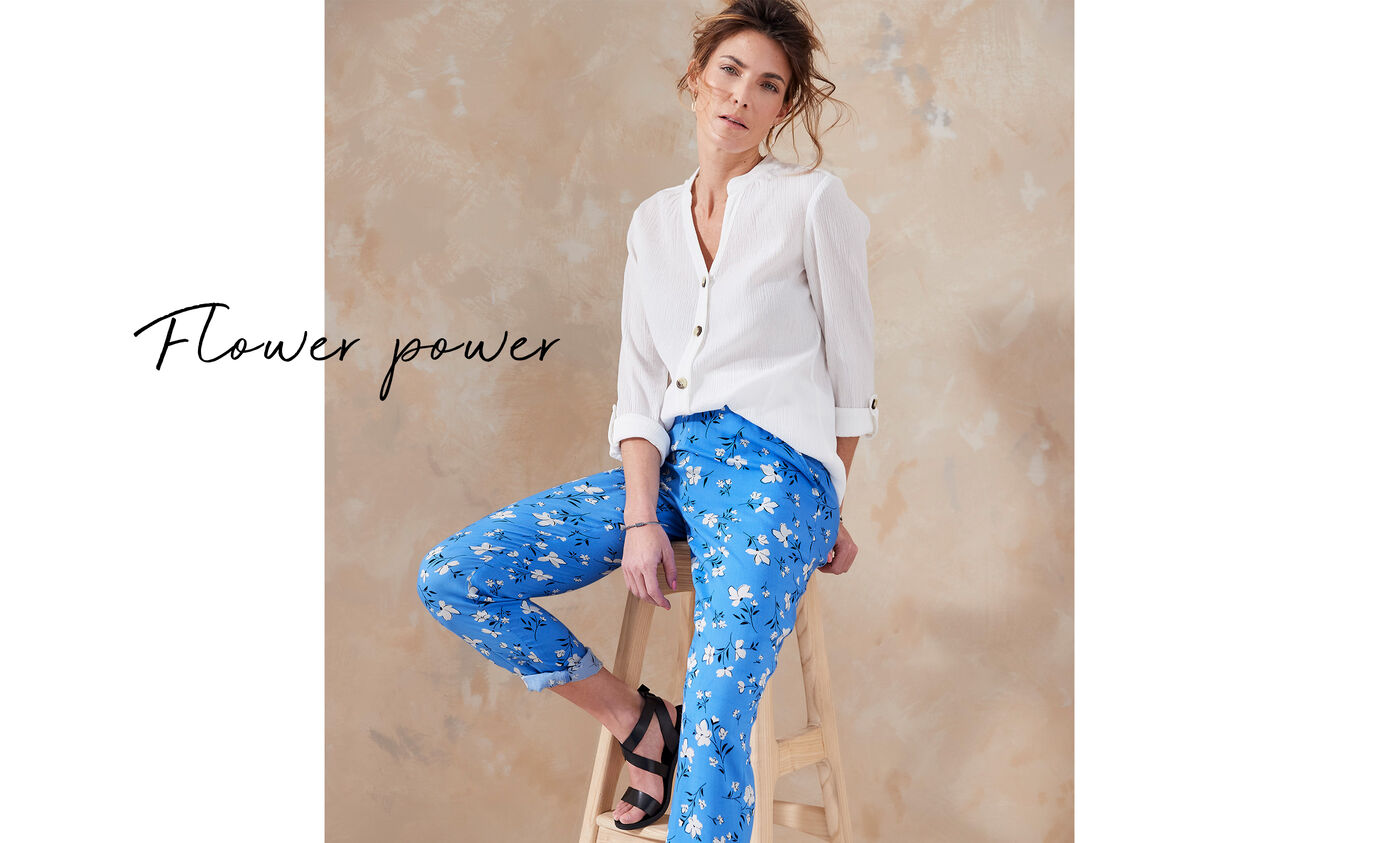 Woman leaning on a chair wearing a Cotton Traders white button up blouse with blue floral trousers. It's accompanied by the title 