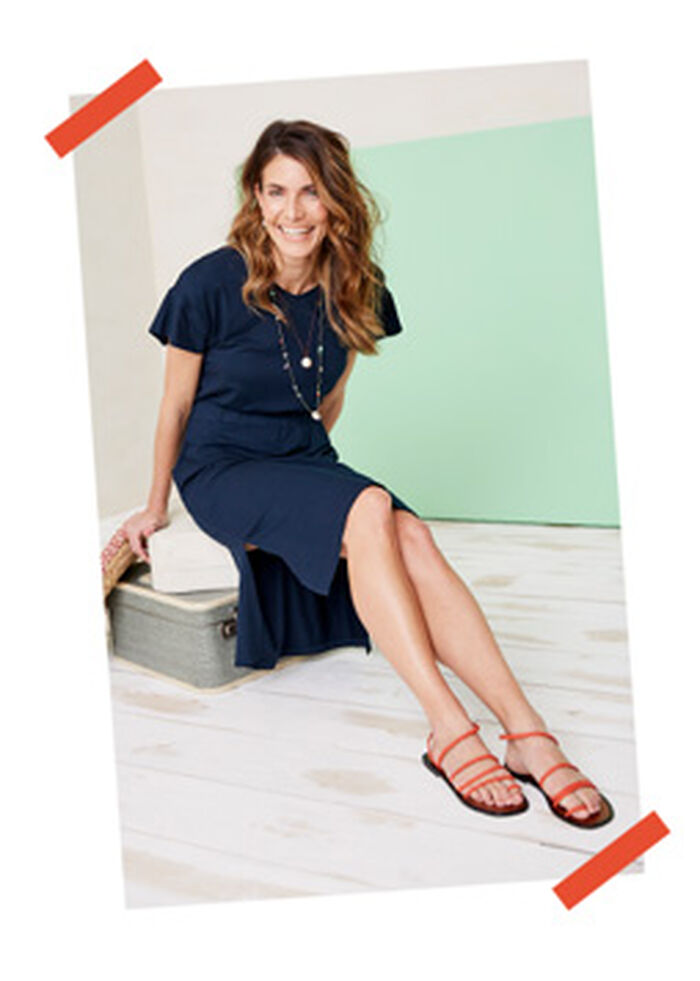 Woman leaning forward while sat on two suitcases. She's wearing a blue Cotton Traders frill sleeve dress with orange sandles and a layered necklace. 