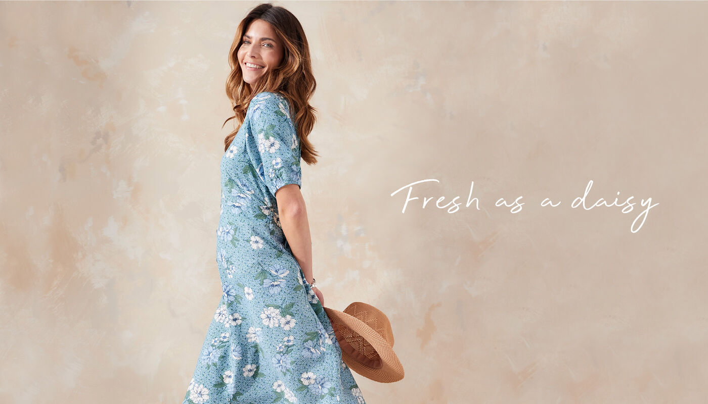 A woman stares off to the side of the camera. She's wearing a blue floral maxi Cotton Traders dress and is holding a sun hat behind her back. The image reads 