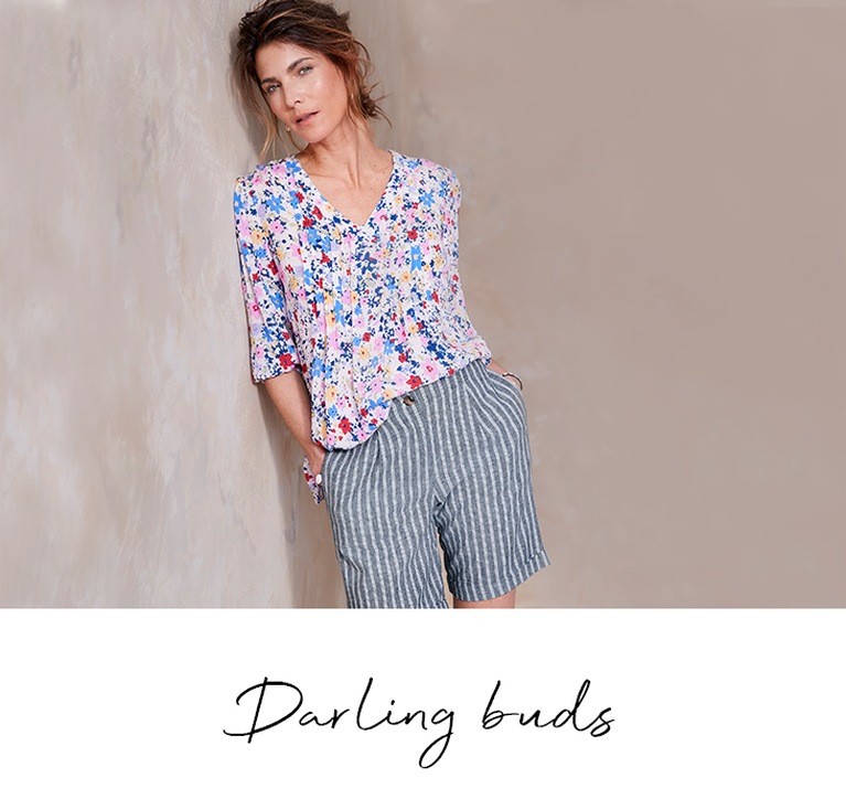 Woman leaning against the wall wearing a Cotton Traders floral blouse and pinstripe shorts with the heading 'Darling Buds'. 