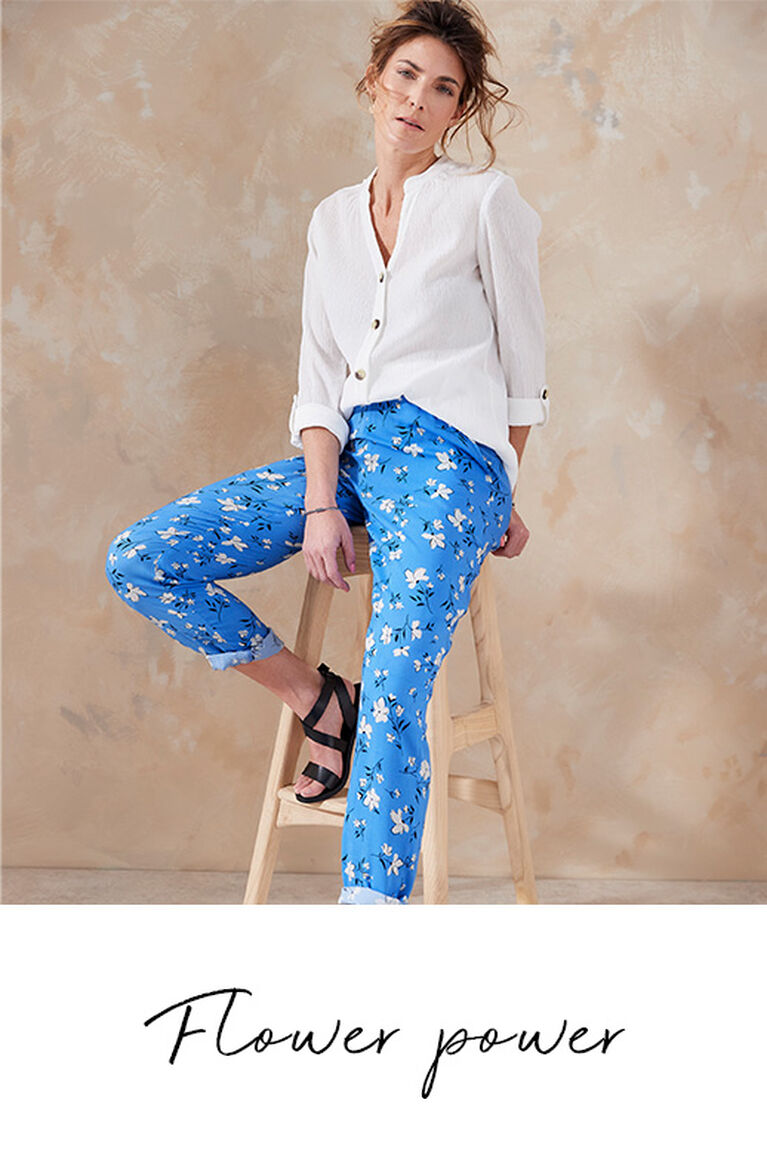 Woman leaning on a chair wearing a Cotton Traders white button up blouse with blue floral trousers. It's accompanied by the title 