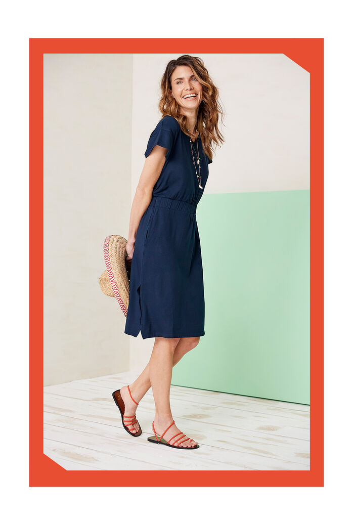 A woman is laughing whilst looking off to the side. She's holding a sun sat behind her and it wearing blue midi dress, layered necklace and Cotton Traders orange sandles. 